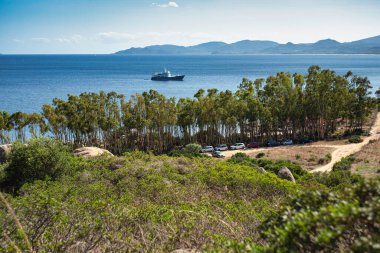 Capo Ferrato 'nun berrak mavi sularında süzülen bir yatın manzarası, sardunya, yemyeşil bitki örtüsü ve ön planda park etmiş arabalar.