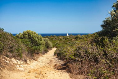 Güneşli bir günde İtalya 'nın Sardunya kentindeki Capo Ferrato sahilinde beyaz bir deniz fenerine doğru giden toprak yolun manzarası.