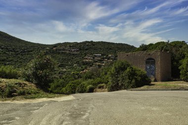 Ön planda terk edilmiş bir bina var. Arka planda yemyeşil bir tepe köyü var. Kosta Rei, Sardunya 'da canlı mavi bir gökyüzünün altında.
