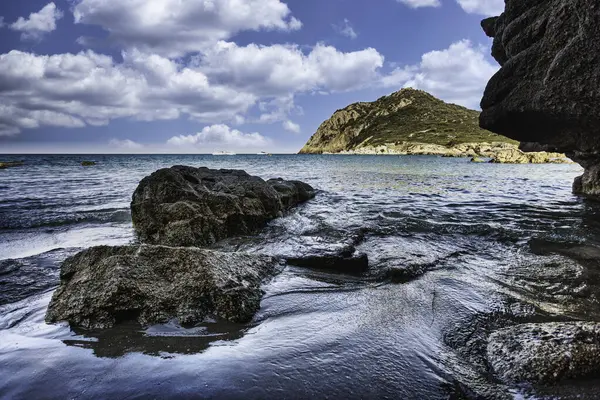 İtalya, Sardunya 'daki manzaralı Cala sa figu plajında kayaların üzerinde hafif dalgalar, açık mavi bir gökyüzü ve arka planda bir tepe.