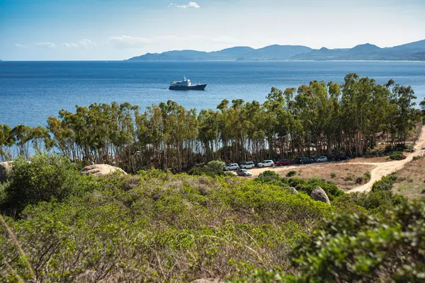 Capo Ferrato 'nun berrak mavi sularında süzülen bir yatın manzarası, sardunya, yemyeşil bitki örtüsü ve ön planda park etmiş arabalar.
