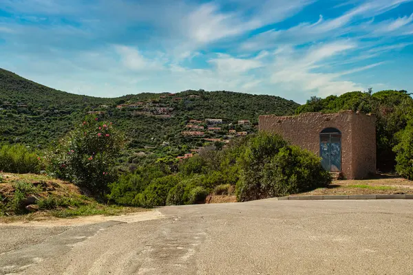 Ön planda terk edilmiş bir bina var. Arka planda yemyeşil bir tepe köyü var. Kosta Rei, Sardunya 'da canlı mavi bir gökyüzünün altında.