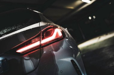 Car taillight glowing red in a dark parking garage, modern led pattern reflecting on wet vehicle surface with rain droplets and moody urban bokeh atmosphere