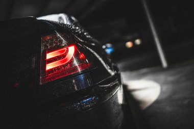 Dark car taillight glowing red through raindrops on a wet surface in a moody parking garage at night, reflecting urban lights and textured droplets on the vehicle rear