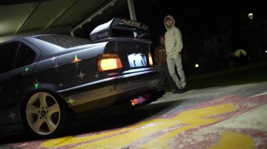 Raindrops falling at night, illuminated by the bright light of a modern street lamp against a dark sky, with green tree leaves framing the moody and atmospheric scene of a summer storm