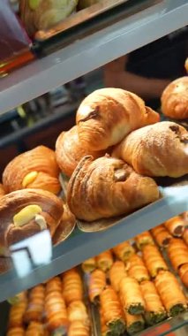 Freshly baked italian croissants and cannoli with various fillings like cream, chocolate, and pistachio resting on trays inside a refrigerated glass display case at a local pastry shop