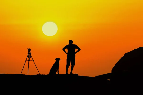 A man and his dog stand on a hill, silhouetted against the sky, tripod stands beside them, set up for photography. The man and his loyal companion embrace the beauty of nature together