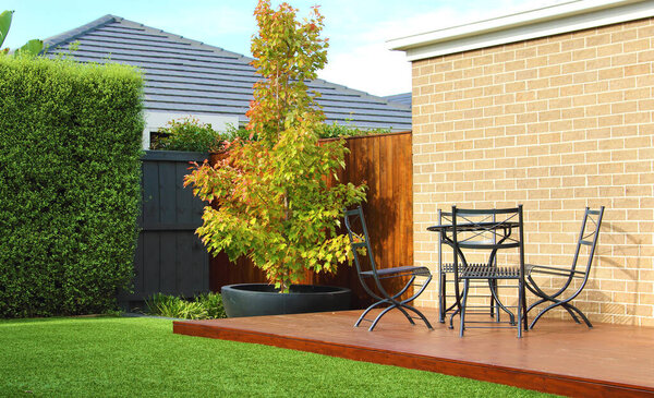 Australian backyard showcases a lush artificial grass lawn and a majestic tree in the background, providing a natural touch against a wooden fence, wooden decking area furnished with stylish outdoor furniture, all set against brick wall