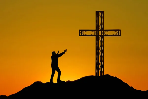 A lone figure stands before a cross perched on a mountain, his hands raised in prayer