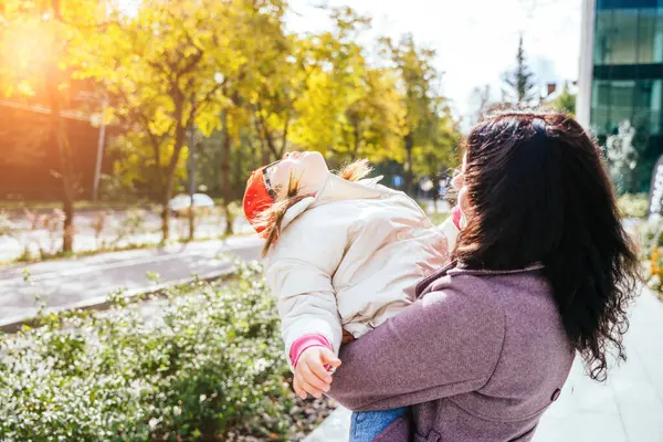 Cute lovely positive brunette mother carrying her happy disabilities daughter. Woman and child enjoy walk autumn sunny day outside. Sun glare effect.