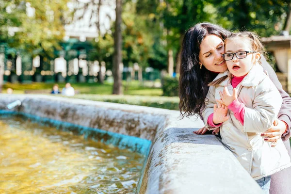 特別なニーズを持つ小さな女の子は 秋の公園の屋外で母親と過ごす時間を楽しんでいます 子供は親からの愛を感じます 噴水の近くに娘と幸せな女性の肖像画 — ストック写真