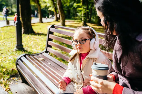 Special need child wearing headphones and happy time to use a smartphone on bench with her mother in park outdoor.