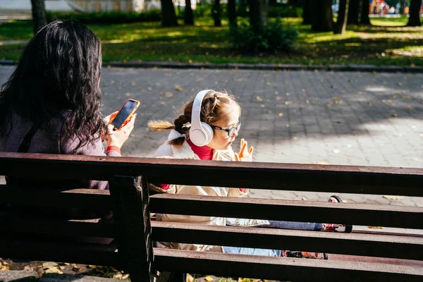 Special need child wearing headphones and happy time to use a smartphone on bench with her mother in sunny park outdoor.