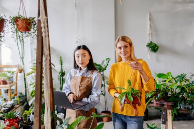 Sarı kazaklı mutlu bir orta yaşlı kadın elinde çiçekle elinde Asyalı kadın satıcıyla fabrikanın iç kısımlarında dizüstü bilgisayar tesisleri tutuyor....
