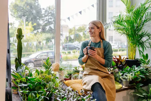 商界女性店主在花店里轻松地喝咖啡 在橱窗里被植物包围 看上去很积极 不怕挑战 创业的概念 — 图库照片