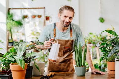 Önlük takan yakışıklı genç adam dikkatli bir şekilde saksıdaki çiçekleri suyla dolduruyor. Bitki bakımı ve ev bahçesi kavramı. Küçük işletme girişimcisi. Şehir ormanı ve bitki ebeveyni kavramı.