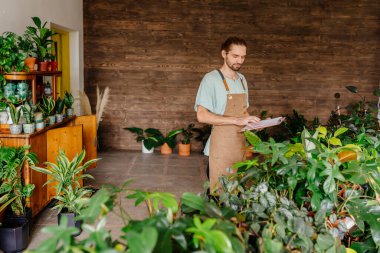 Çiçekçide çalışan başarılı yakışıklı erkek çiçekçi önlük giyen bitkilerin listesini çıkarıyor. Çiçekçide erkek çiçekçi. Çiçekçilik ve insanların konsepti.