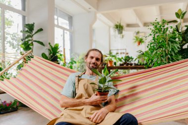 Gülümseyen bahçıvan hamakta rahatlıyor. Bahçenin ortasında Ficus 'u tutarak rahatlıyor. Evde yeşillik var. Bitki sevgisi. Kapalı bir bahçe..