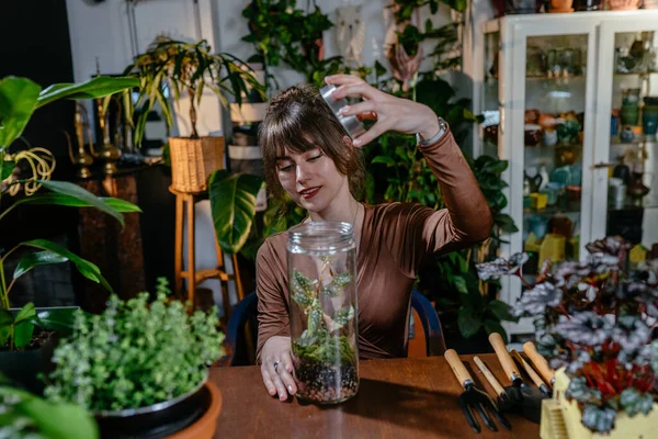 Girl seller in a plant store in her workplace takes care of a plant in a jar removes dry leaves with tweezers.