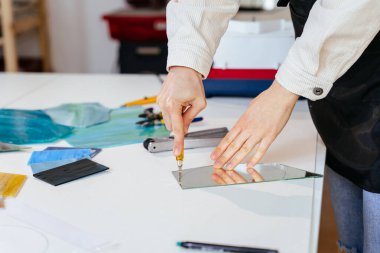 Kadın ellerini kapatarak atölyesinde gelecekte vitray olacak bir ayna parçası hazırlıyor. Lekeli cam sanatı, cam işçiliği.