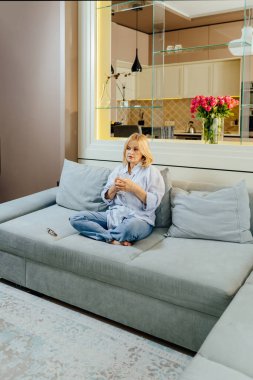 Düşünceli, yorgun, üzgün, olgun, orta yaşlı bir kadının Lotus 'ta koçun üzerinde oturduğu oturma odasının iç kısmında poz verdiği dikey bir fotoğraf. Kapalı alan, insanların kavramı.