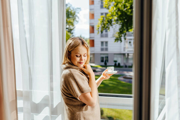 Charming lovely mature 55s blond female standing in front of window under sun rays, drinking coffee or tea in morning, dreaming imagining their future.