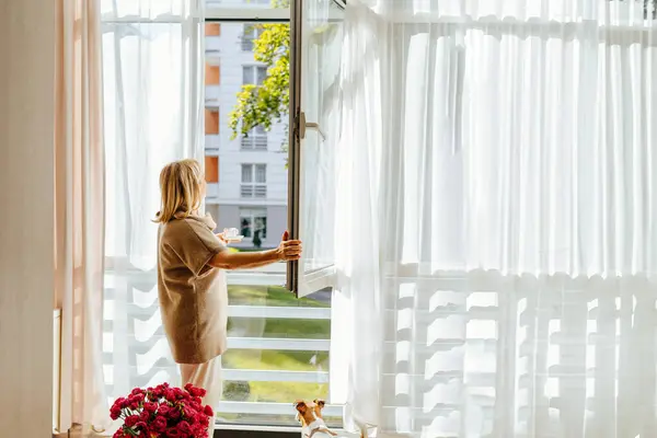 Woman having cup of coffee open the window, breathing fresh air, enjoy sunny day, looking outside.