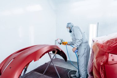 Koruyucu giysiler içinde erkek araba boyacısı ve maske takan kırmızı boyalı araba tamponu ve oda atölyesinde vernik..
