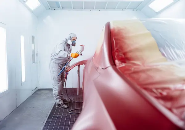 Mechanic in Painting Booth spray a car in chamber. Car service station. Worker painting a red electric car in special garage, wearing costume and protective gear.