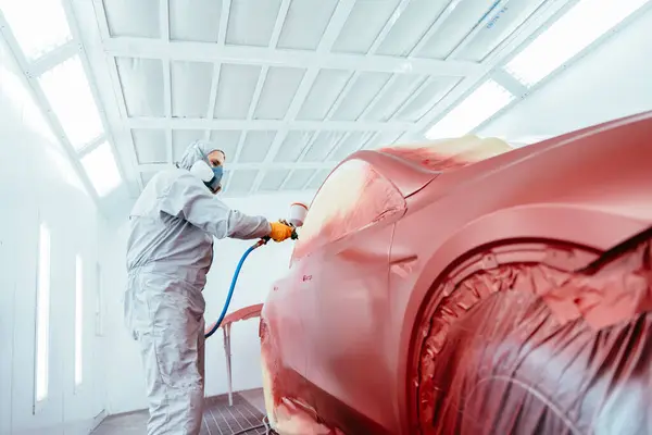 Person spray painting car in red color in special car chamber. Car service station. Worker painting a red electric car in special garage, wearing costume and protective gear.