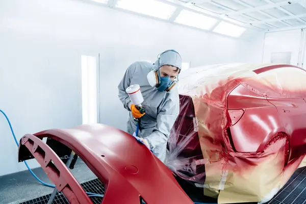 Male car painter in protective clothes and mask painting automobile bumper with red paint and varnish in chamber workshop.