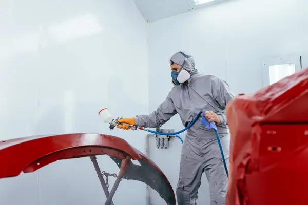 Authentic shot. Car painter in protective clothes and mask painting automobile bumper with metallic paint and varnish in chamber workshop.