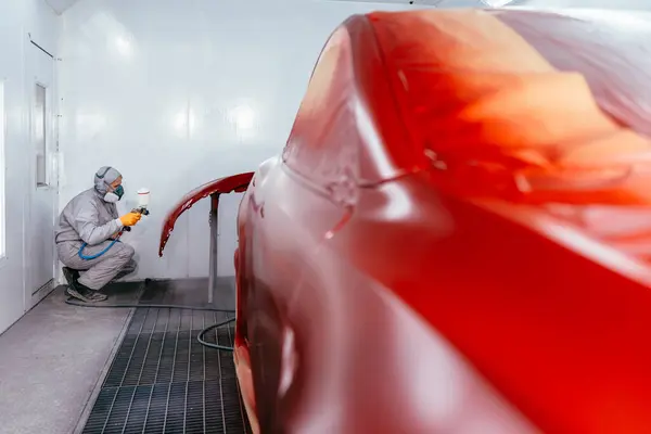 Professional male car mechanic working with spray gun painting part of auto body in automotive paint service shop. Automobile painter painting car in chamber.