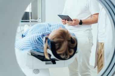 Bir kadın hastane odasında bir yatakta yatıyor ve arkasında bir adam var. Adam bir tablet tutuyor ve kadına bakıyor. Kadın çizgili bir gömlek giyiyor ve başı aşağıda.