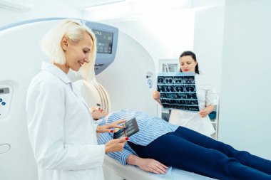 Arka plan hemşiresi hastayı muayene testine hazırlarken, MRI ekranındaki röntgen görüntülerini inceleyen kadın doktor.
