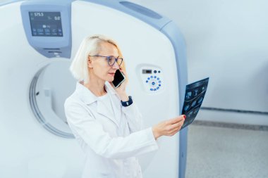 Modern tomografi odasındaki neşeli sarışın kadın doktor portresi telefon görüşmesi yapıyor ve arka planda CT tarayıcısıyla röntgen görüntüsü arıyor..