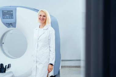 Beyaz önlüklü bir kadın tıbbi görüntüleme makinesinin önünde duruyor. Gülümsüyor ve mutlu.