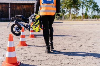 Güvenlik yeleği giymiş biri elinde motosiklet kaskıyla otoparktan geçiyor. Sahne, motorsiklet sürücüsü ya da motorsikletle ilgili bir alanda çalışan olabileceğini gösteriyor.