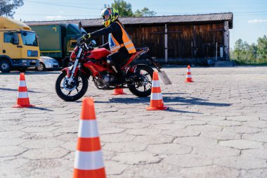 Otoparkta turuncu ve beyaz külahlı bir adam motosiklet sürüyor. Kozalaklar sıraya dizildi ve adam kask takıyor.