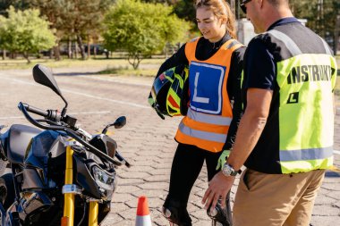 Motosiklet kasklı, L harfli bir kadın yelekli bir adamdan talimat alıyor. Öğrenci ve öğretmen, motosiklet okulunda sınav..