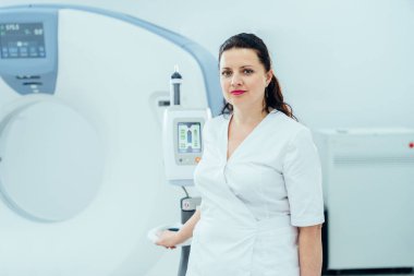 Kendine güvenen kadın radyoloji uzmanı ve MRI tarayıcısı. Beyaz laboratuvar önlüklü kadın tomografinin yanında duruyor..