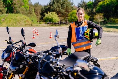 Güvenlik yeleği ve kask takan bir kadın bir motorsikletin yanında duruyor. Kadın gülümsüyor ve poz veriyor..