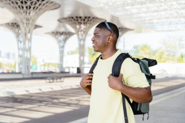 Sarı tişört ve sırt çantası giyen siyah bir adam şehrin arka planında duruyor. Gülümsüyor ve mutlu.