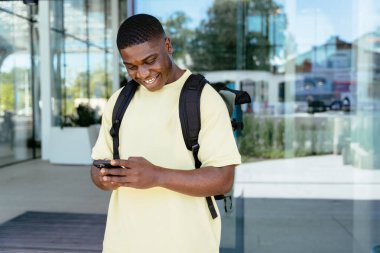 Genç Afrikalı bir adam gülümsüyor ve cep telefonuna bakıyor. Üzerinde sarı bir gömlek ve açık hava terminalinin sırt çantası var. Seyahat, macera, rezervasyon, hedef, konsept.