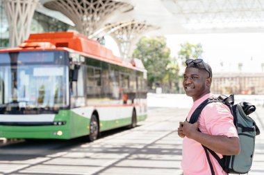 Sırt çantalı Afrikalı bir adam otobüs durağında yeşil beyaz bir otobüsün önünde duruyor..