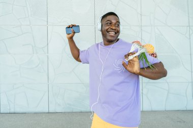 Kulaklıkla müzik dinleyen mutlu Afrikalı bir adam açık hava istasyonunda market alışverişi yapıyor. Millennial etnik adam elinde taze sebzelerle ağ torbası tutuyor. Sürdürülebilir yaşam tarzı kavramı.