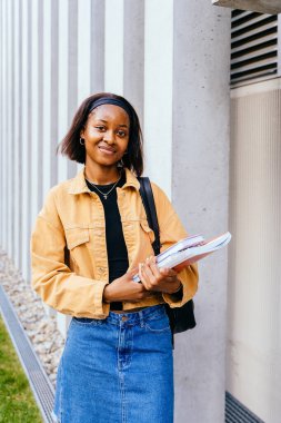 Sarı ceket ve kot pantolon giyen genç bir kadın bir yığın kitap tutuyor. Yüzünde bir gülümseme var ve mutlu.