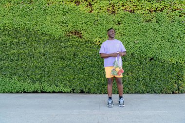 Arka planda yeşil bitki duvarı olan meyve ve sebze dolu sürdürülebilir market poşeti kullanan mutlu bir Afrikalı. Çevre dostu ağ tüccarı. Sıfır atık, plastiksiz konsept