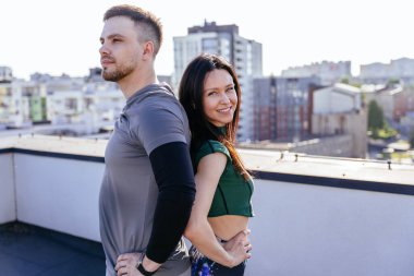 Atletizm kıyafetleri giymiş sevimli bir çift arka planda çatıda sırt sırta duruyorlar. İkisi de omuzlarının üzerinden bakıyor.