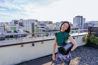 Spor salonu rutin gereksinimleri. Orta yaşlarda aktif ve sağlıklı yaşam biçimi kavramı. Güzel esmer seksi kadın şık spor kıyafetleriyle çatıda yoga minderi tutuyor şehir manzaralı..